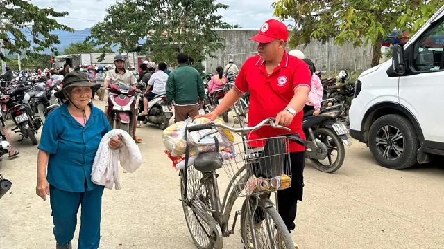 Tình nguyện viên Chữ thập đỏ dắt xe đạp chở quà đi cạnh một cụ bà
