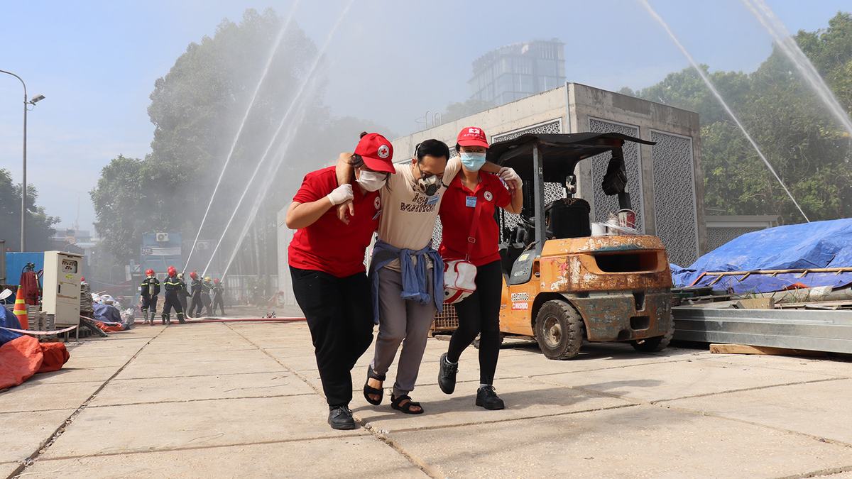 Hai tình nguyện viên Chữ thập đỏ đội mũ đỏ dìu một người đeo mặt nạ phòng độc ra khỏi khu vực giả định nguy hiểm, phía sau có vòi nước phun