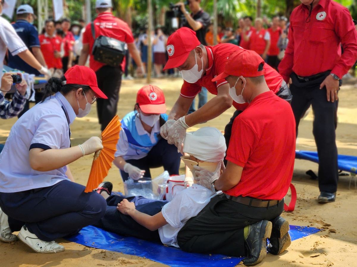 Tình nguyện viên Chữ thập đỏ đội mũ đỏ hướng dẫn băng bó đầu cho một người nằm trên tấm bạt xanh trong buổi thực hành sơ cấp cứu