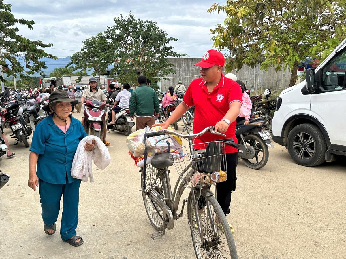 Tình nguyện viên Chữ thập đỏ áo đỏ dắt chiếc xe đạp chở túi quà, đi cạnh một cụ bà giữa đông người dân
